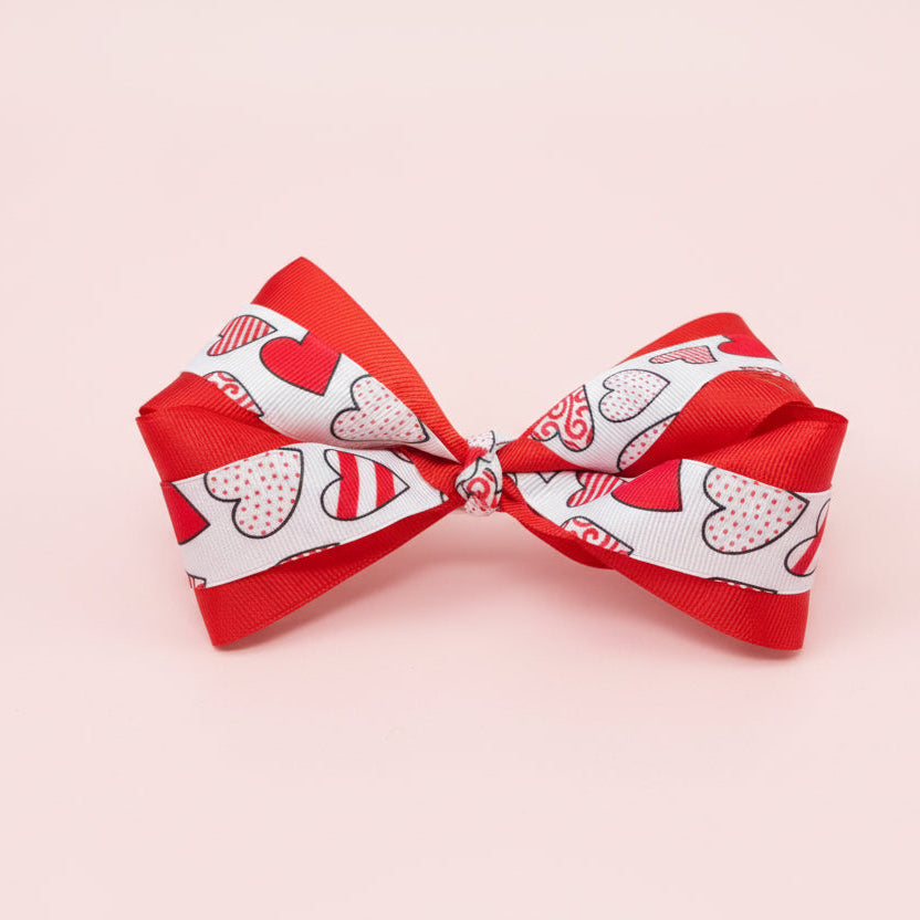 Red layered grosgrain hair bow with a white ribbon overlay featuring red and pink heart patterns, photographed on a pale pink background.
