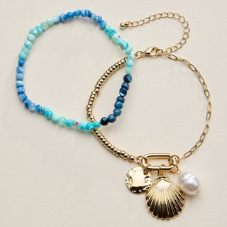 Two bracelets on a pink background, one with blue beads and the other with gold chain and shell charm.