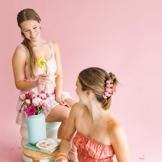 Two women sitting on a pink background with a small table between them, one holding flowers.