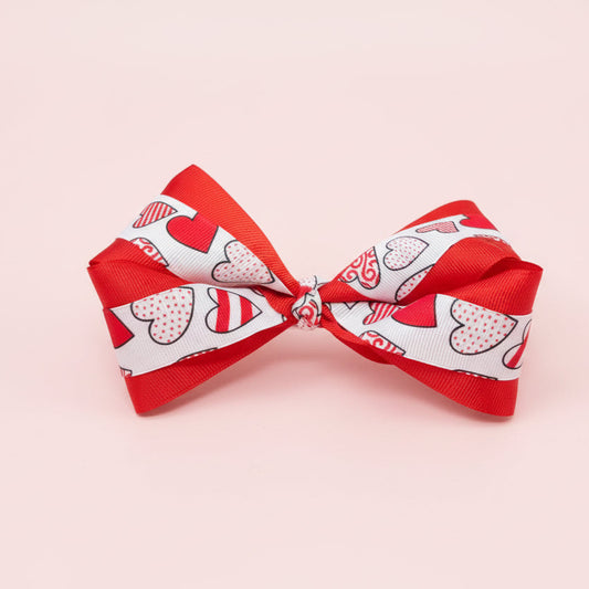 Red layered grosgrain hair bow with a white ribbon overlay featuring red and pink heart patterns, photographed on a pale pink background.