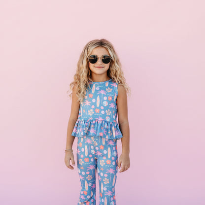 Young girl in a blue floral jumpsuit standing in front of a pink background. 