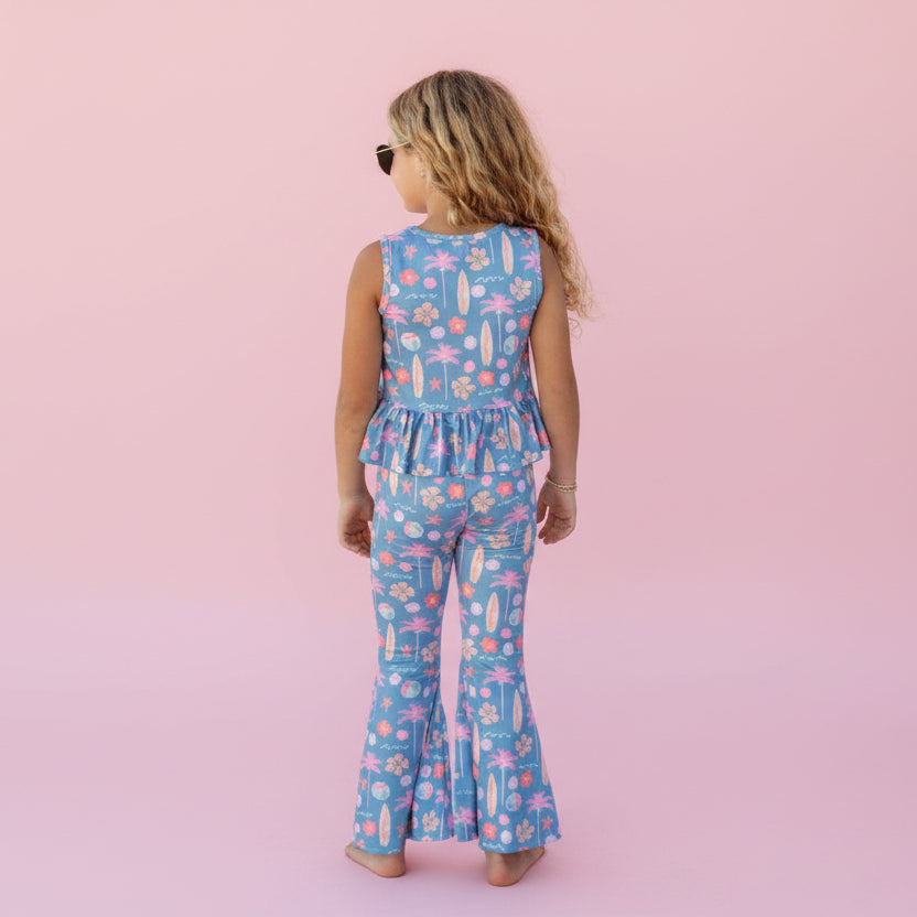 Child wearing a blue floral jumpsuit standing in front of a pink background. .