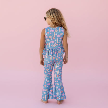 Child wearing a blue floral jumpsuit standing in front of a pink background. .