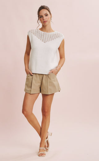 Woman wearing a white top with cut-out details and beige shorts on a pink background