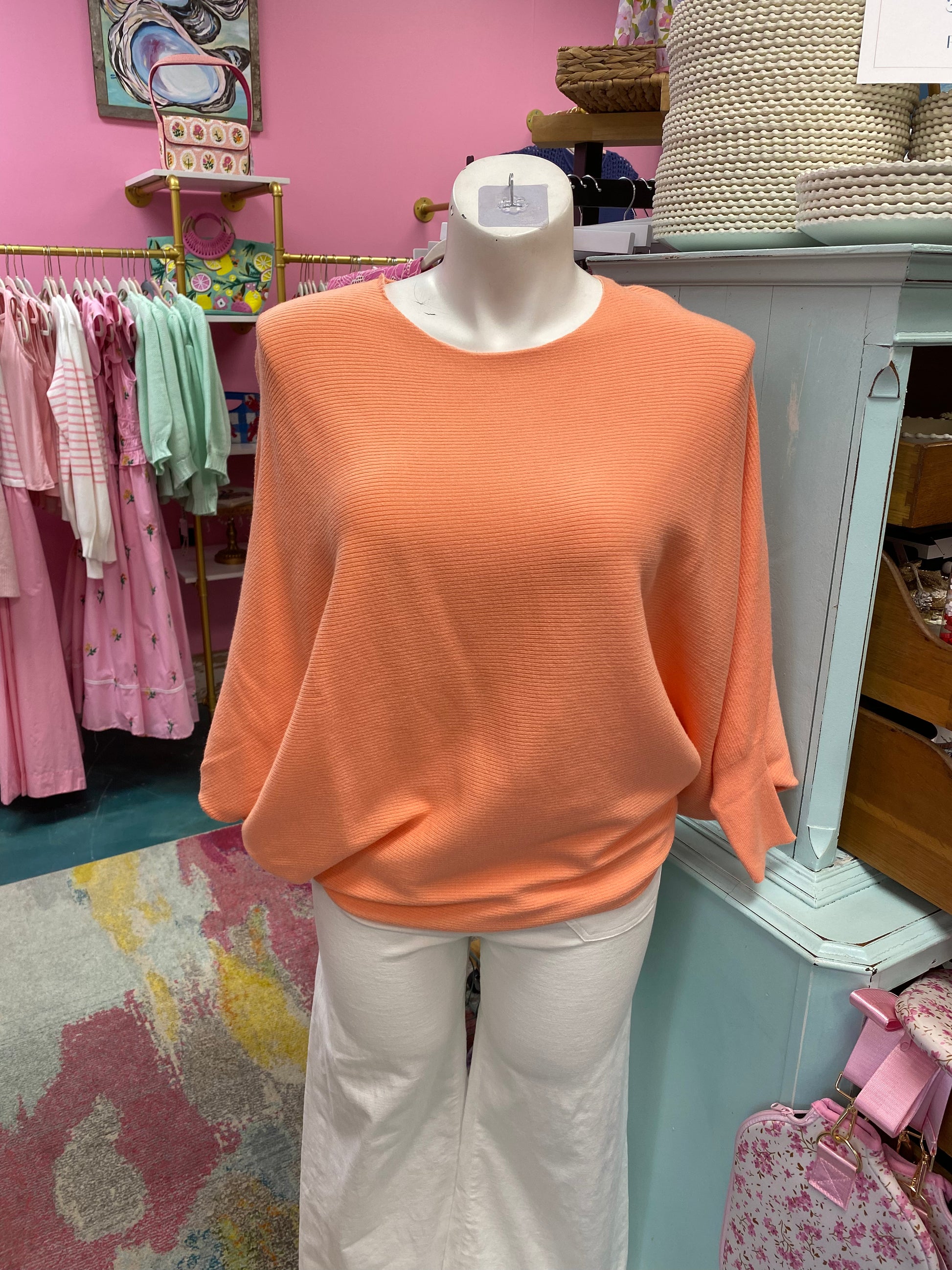 Mannequin wearing an orange sweater and white pants in a store setting with pink walls and clothing racks.
