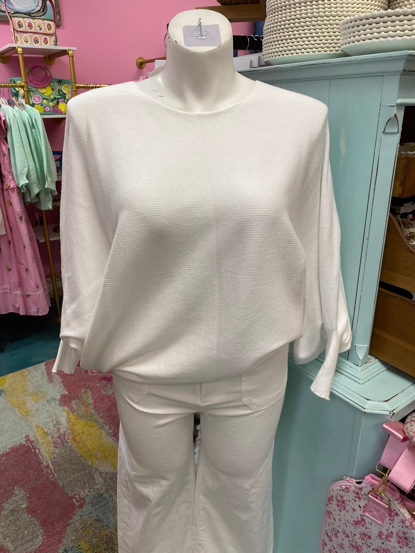 White sweater on a mannequin in a store setting with pink walls and various items in the background.